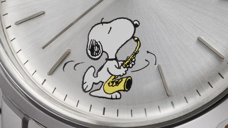 A close-up of a wristwatch dial featuring a silver sunburst background with minimalist hour markers. The design includes an illustration of Snoopy from the Peanuts comic strip, joyfully playing a yellow saxophone. The whimsical graphic is placed near the lower-left quadrant of the dial, adding a playful and nostalgic touch to the watch’s otherwise sleek and refined design.