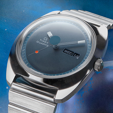 A close-up view of the Q Timex 1975 Enigma Reissue watch, showcasing its sleek brushed stainless steel case and integrated bracelet. The dial features a deep blue gradient finish with minimalist hour markers, a day-date window at 3 o'clock, bold rectangular hands, and a signature orange dot at the 8 o'clock position, all set against a cosmic blue background.