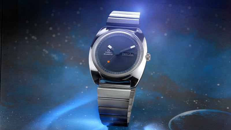 A sharp close-up of the Q Timex 1975 Enigma Reissue watch dial, highlighting the clean typography and retro-modern aesthetic. The dark blue face features white rectangular indices, prominent angular hands, and a day-date window showing “WED 14.” The vibrant red-tipped seconds hand stands out against the minimalist backdrop.