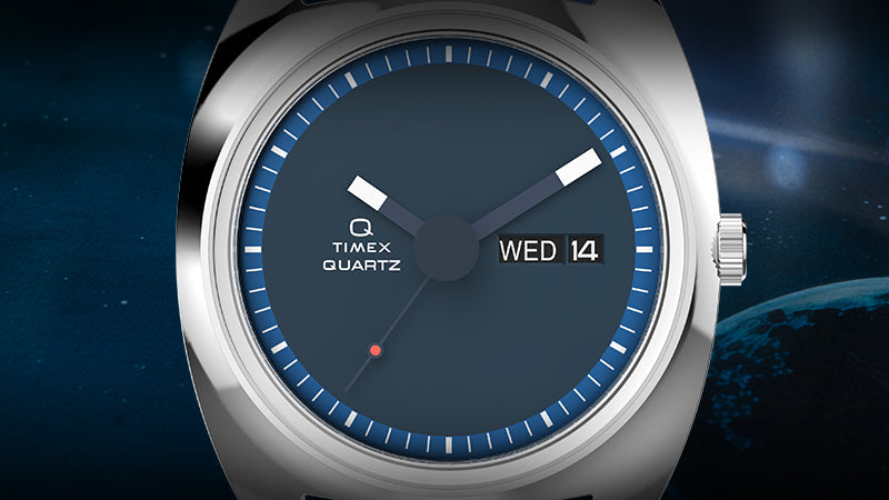 A front-facing full view of the Q Timex 1975 Enigma Reissue watch suspended in a starry, cosmic blue environment. The timepiece displays its brushed stainless steel case and band, minimalist deep blue dial with bold hour and minute hands, a day-date display at 3 o’clock, and a single orange dot near 8 o’clock for a signature accent.
