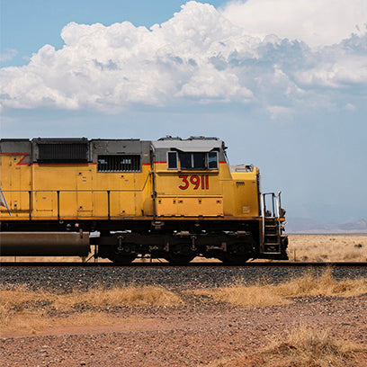 A bright yellow locomotive marked with the number 3911 travels across a dry, open landscape under a partly cloudy sky. The image captures the spirit of American railroads and industrial movement.