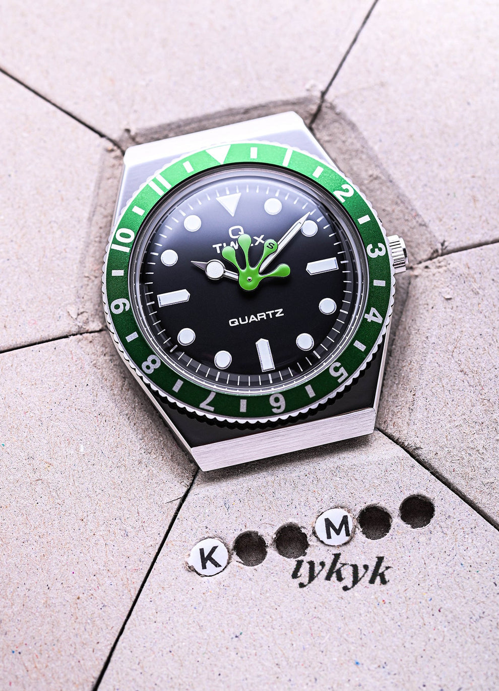 Timex x seconde/seconde/ watch placed on light grey hexagonal tiles. The black dial with green frog hand and green-and-black bezel is accompanied by playful typography reading 'K M iykyk' made of punched-out paper dots.