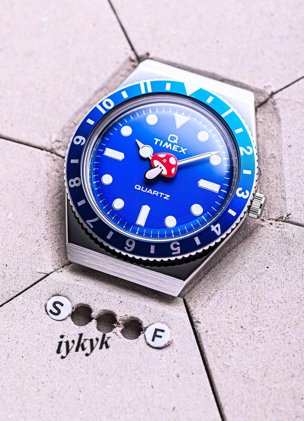 Flatlay of the Timex x seconde/seconde/ Episode 0 watch resting on pale concrete tile. The blue bezel and dial pop against the textured surface, with the mushroom hand at the center. Below, punched-out letters spell 'S F iykyk'.