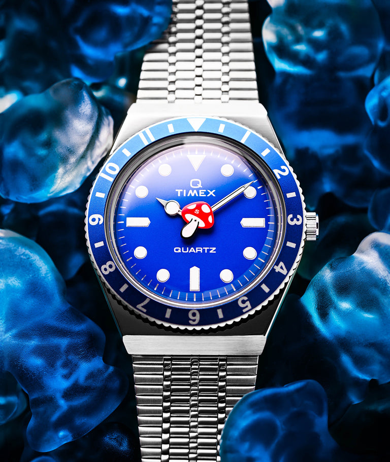 A Timex Q Quartz watch with a vibrant blue dial and bezel, featuring a red mushroom-shaped seconds hand, stainless steel bracelet, and white hour markers. The watch is displayed on a bed of translucent, blue-toned gummy bears.