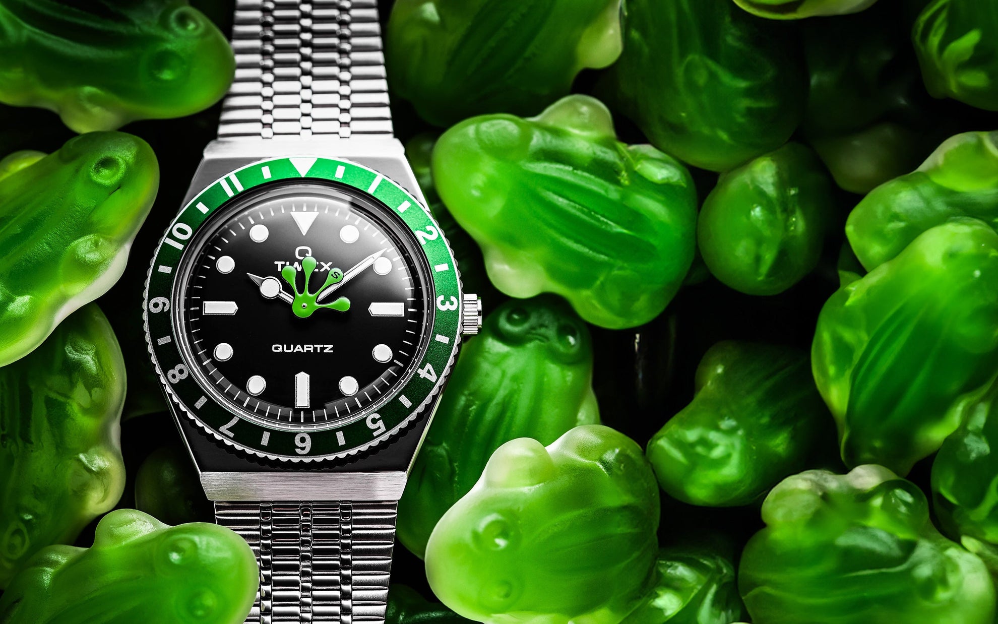 Timex x seconde/seconde/ Q watch with a green and black bezel, black dial, and a playful green frog hand at center. The watch rests on a bed of translucent green gummy frog candies, enhancing the whimsical design.