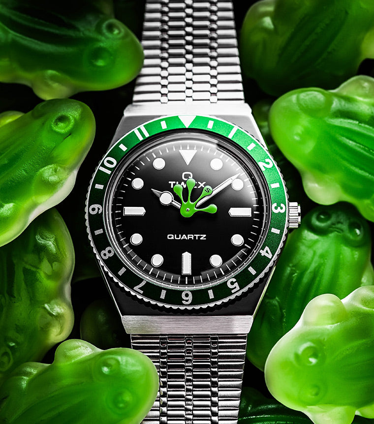 Timex x seconde/seconde/ Q watch with a green and black bezel, black dial, and a playful green frog hand at center. The watch rests on a bed of translucent green gummy frog candies, enhancing the whimsical design.