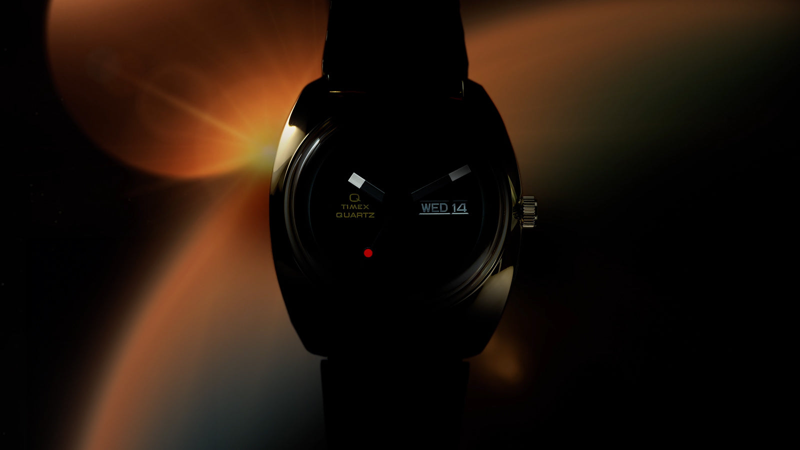 Q Timex® 1975 Enigma watch in a dark, moody lighting setup, highlighting the gold-tone case edges, black dial with geometric hands, red dot detail, and illuminated day-date window, with soft orange light flares in the background.