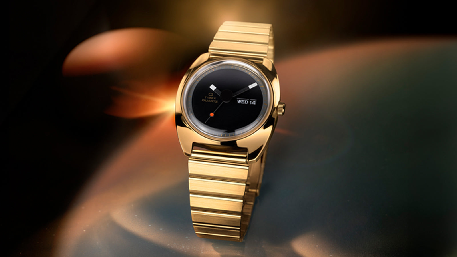 Q Timex® 1975 Enigma watch in polished gold-tone stainless steel, featuring a minimalist black dial with geometric hands, small red dot at 6 o’clock, day-date window at 3 o’clock, and integrated gold-tone bracelet, set against a warm, glowing abstract background.