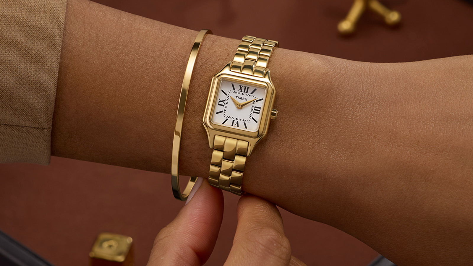 Timex Legacy Addison gold-tone rectangular watch with white Roman numeral dial, worn on the wrist beside a slim gold bangle.