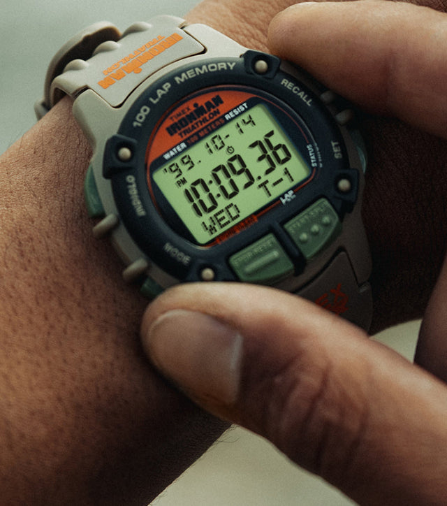 A close-up of a person wearing the TIMEX® IRONMAN® Flix x Huckberry digital sports watch, showing the rugged khaki case, black bezel, and green function buttons. The digital display reads 10:09:36 with the date Wednesday 10-14.