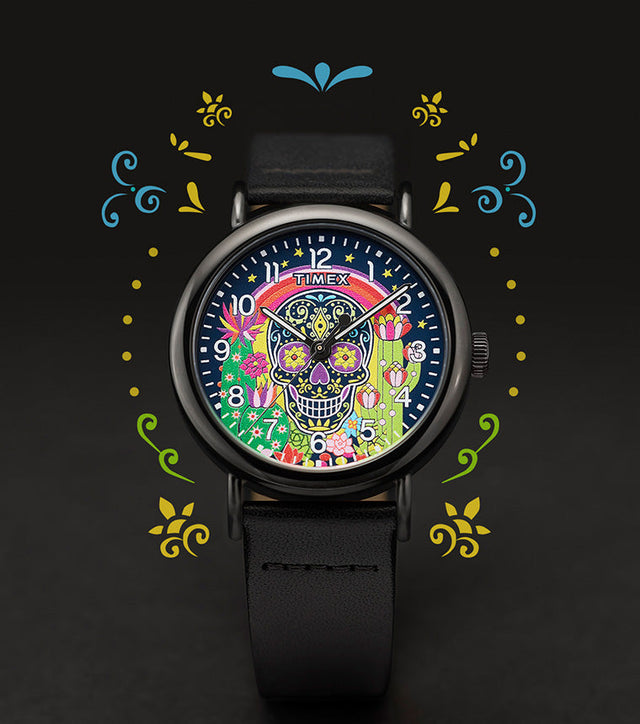 Timex Weekender Day of the Dead watch with a colorful sugar skull, rainbow, stars, and cactus floral design on the dial. The black leather strap is shown with decorative graphics surrounding the watch.