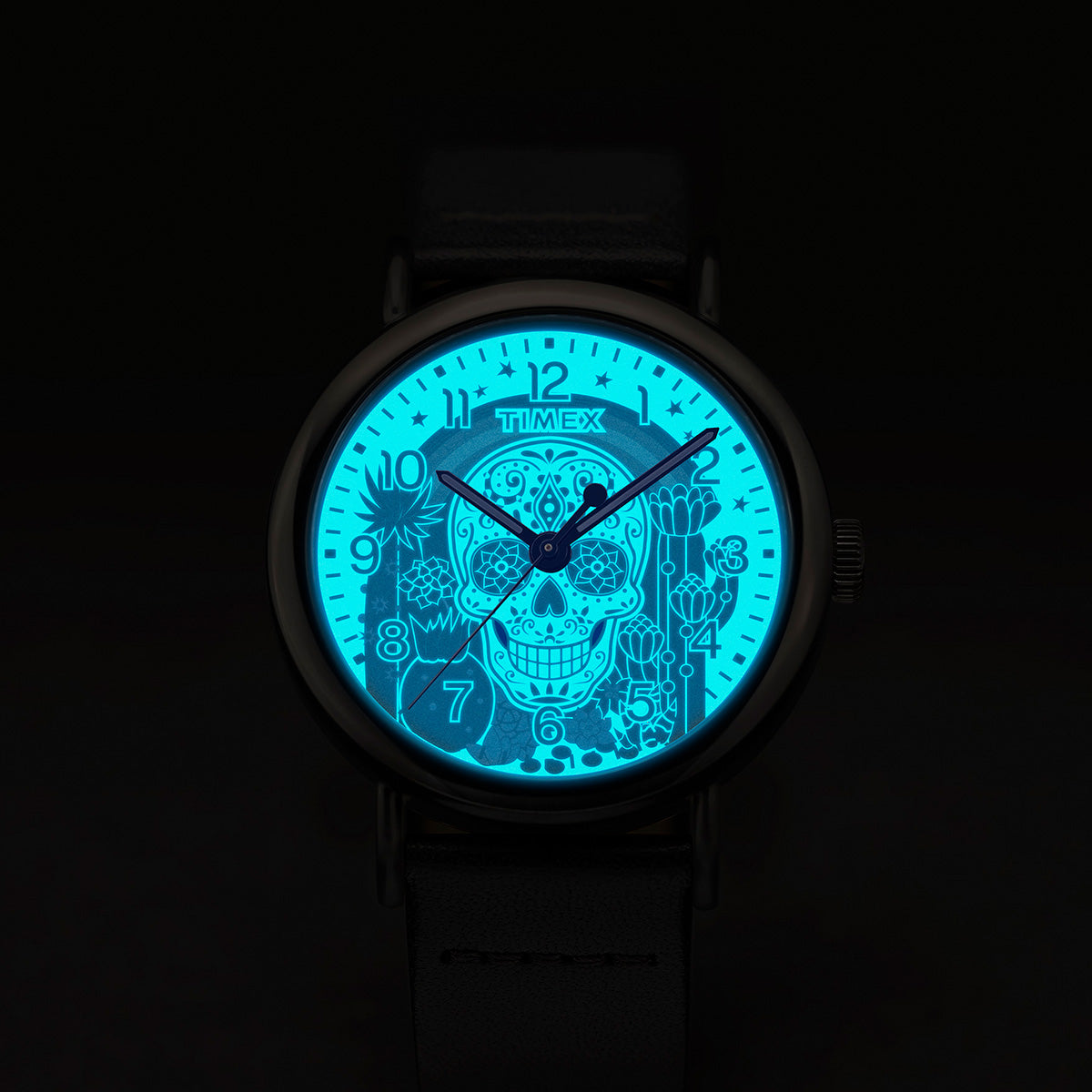 Timex Weekender Day of the Dead watch dial glowing in the dark with INDIGLO® backlight, revealing a bright blue sugar skull and floral details.