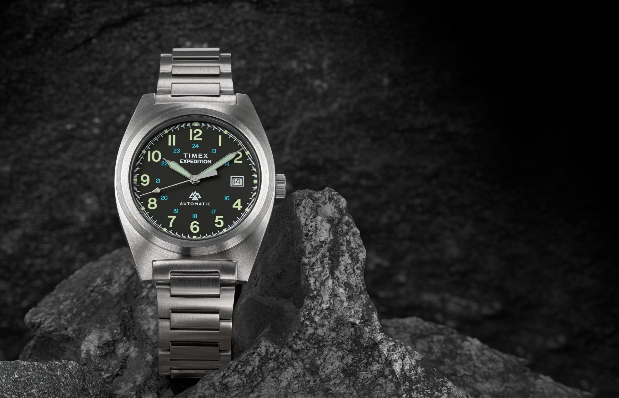 A Timex Expedition® Capstone Automatic watch with a brushed stainless-steel case and bracelet rests among rugged gray rocks. The black dial features luminous green Arabic numerals, a date window at 3 o’clock, and bold hands designed for outdoor durability.
