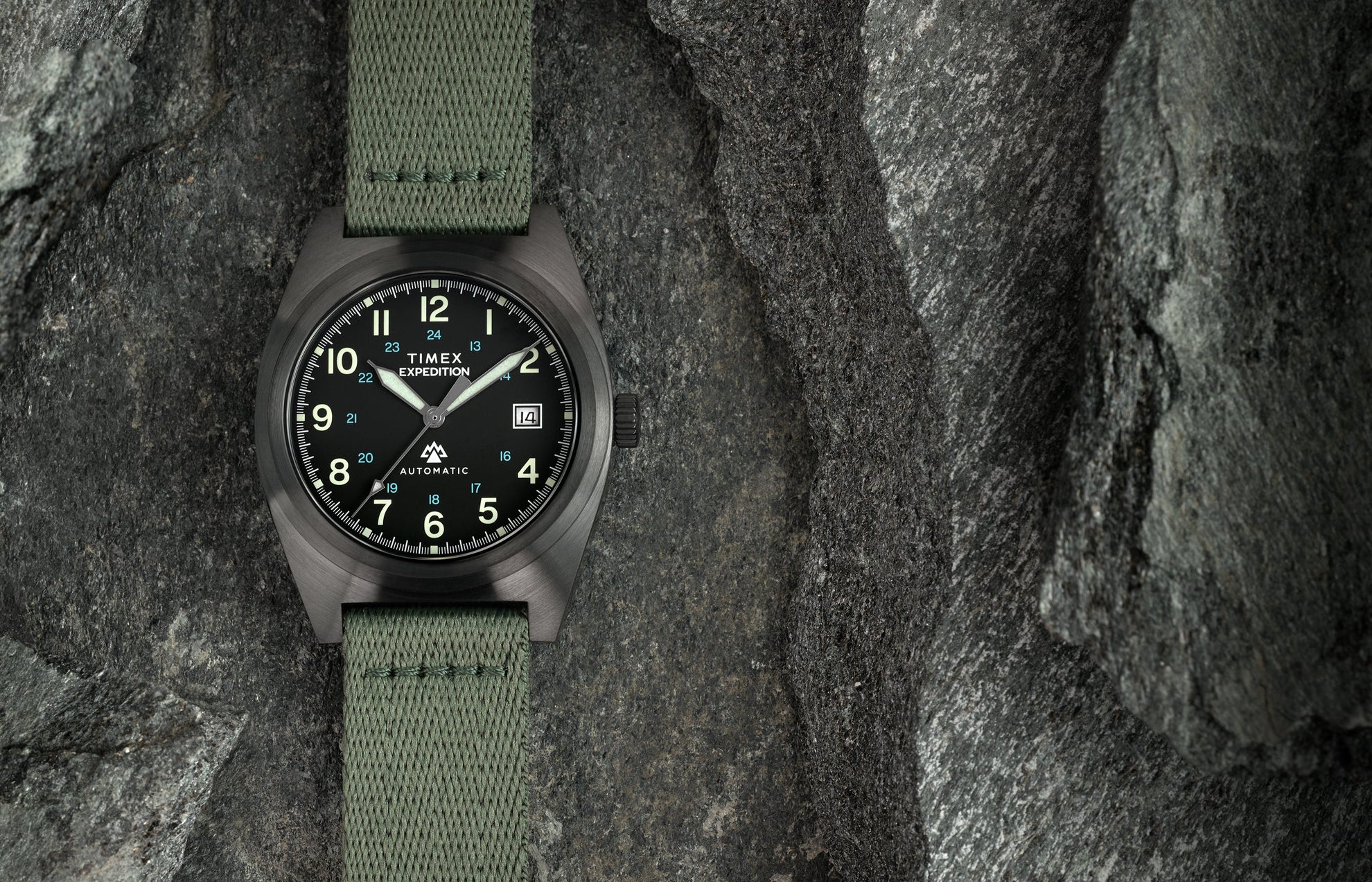 A Timex Expedition® Capstone Automatic watch with a matte gunmetal case and green fabric strap rests among rugged stone surfaces. The black dial features luminous green numerals, blue 24-hour markings, and a date window at 3 o’clock, emphasizing its field-ready durability.