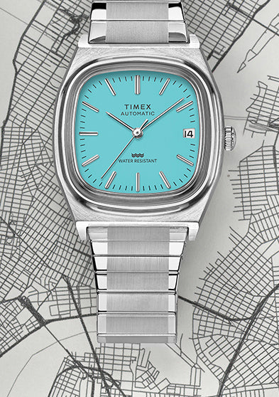 Full view of the Timex® Automatic 1983 E Line Reissue in stainless steel with turquoise dial laid over a grayscale city map. The image emphasizes the retro-inspired square case design and integrated bracelet.