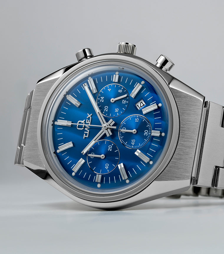 Q Timex Falcon Eye Chronograph with a brushed stainless-steel case and bracelet, featuring a metallic blue dial with three subdials and a date window. The watch is displayed on a light gray background.