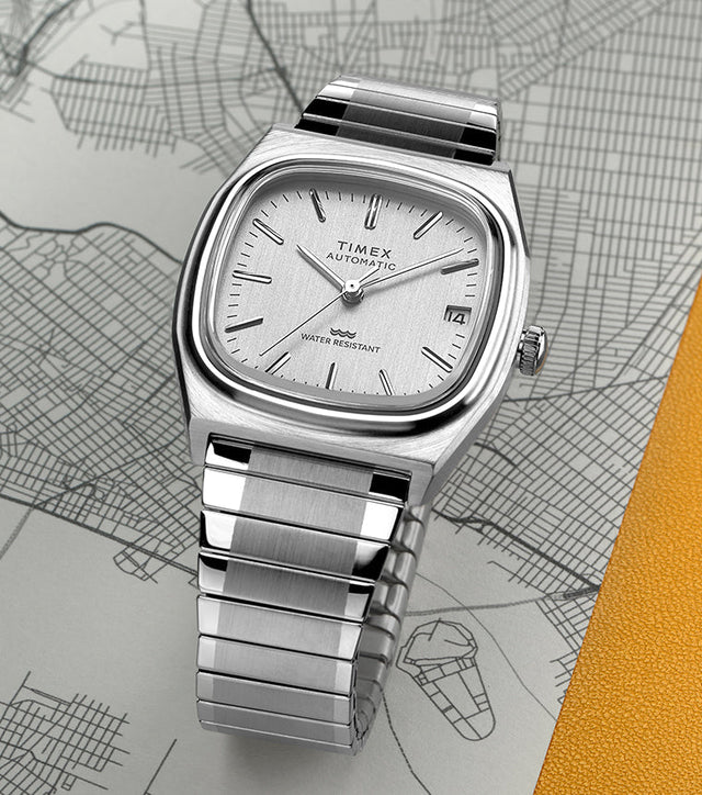 Timex® Automatic 1983 E Line Reissue watch with a brushed stainless-steel case and integrated bracelet, featuring a silver dial with minimalist hour markers and date window at 3 o’clock. The watch is displayed on a city map background with a yellow accent.