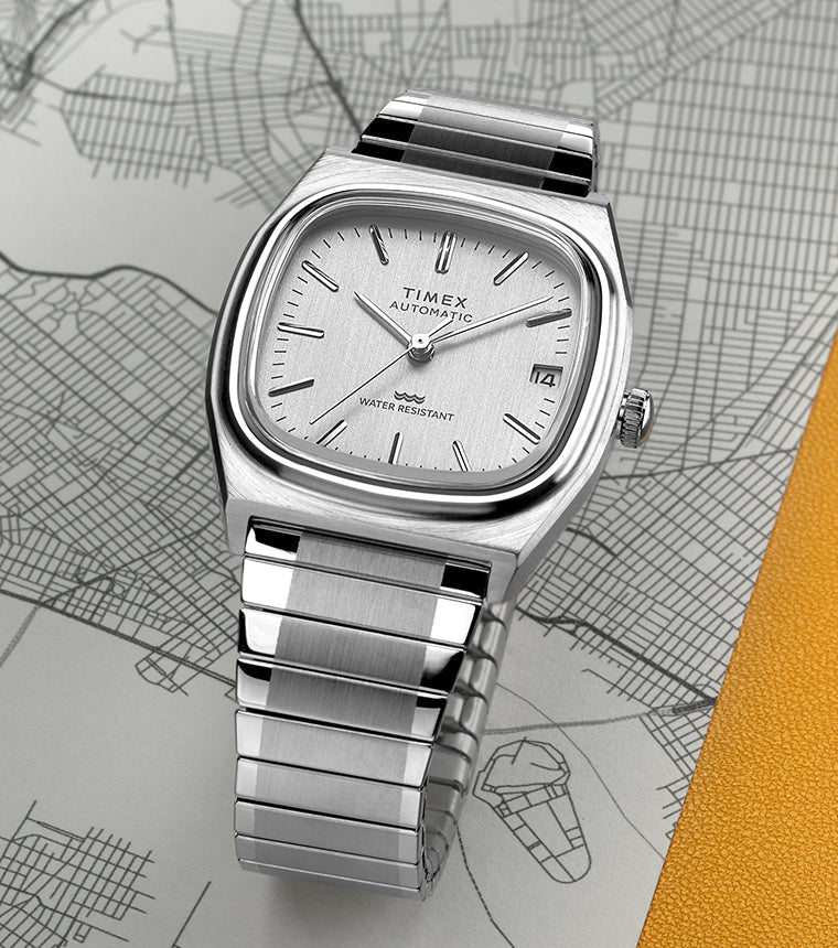 Timex® Automatic 1983 E Line Reissue watch with a brushed stainless-steel case and integrated bracelet, featuring a silver dial with minimalist hour markers and date window at 3 o’clock. The watch is displayed on a city map background with a yellow accent.