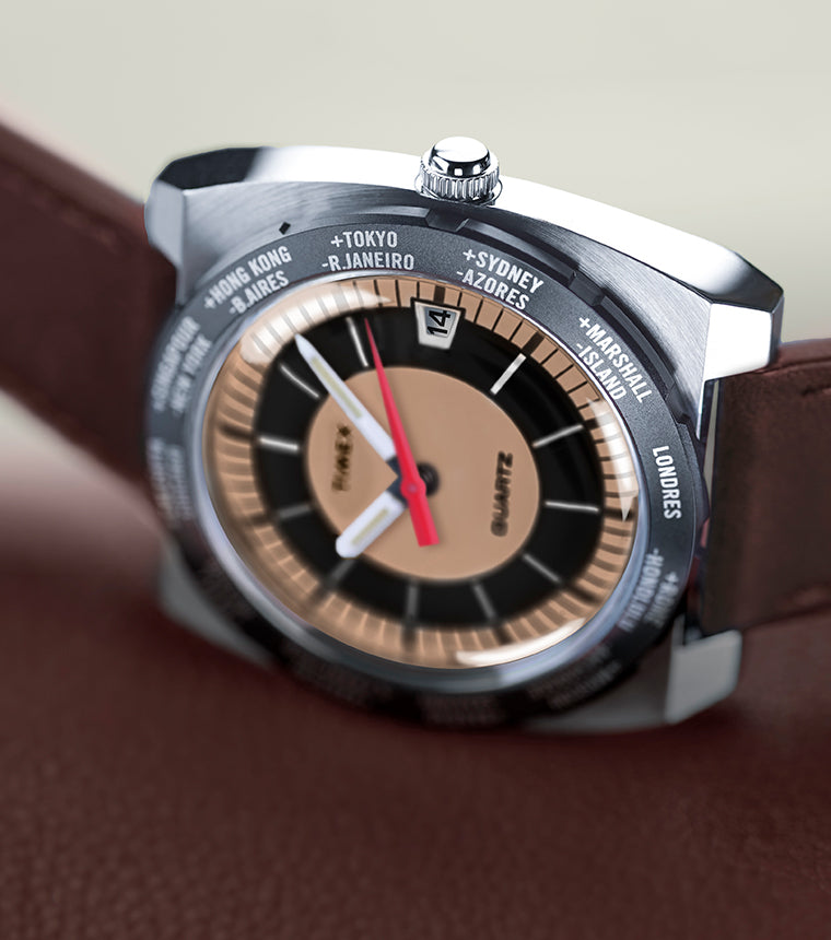 Angled view of the Q Timex® 1972 World Time watch resting on brown leather, showing the brushed stainless-steel case, brown leather strap, and tan-and-black world time dial with a red seconds hand.