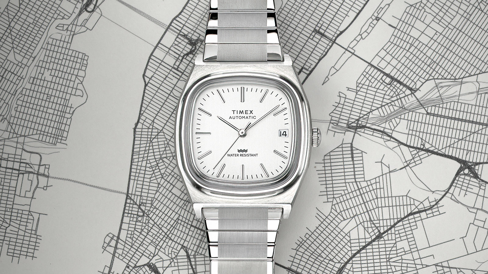 Top-down shot of the Timex® Automatic 1983 E Line Reissue watch lying flat on a map, showing its square cushion-shaped case, silver sunburst dial, and integrated steel bracelet.