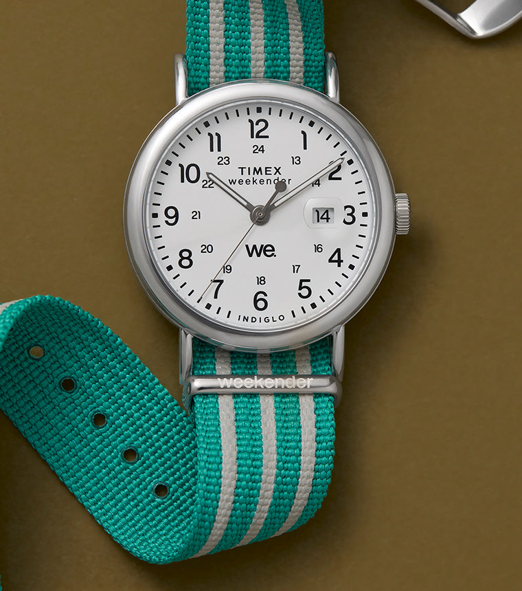 A Timex Weekender watch with a white dial, black numerals, and a teal-and-white striped fabric strap, displayed against a brown background.