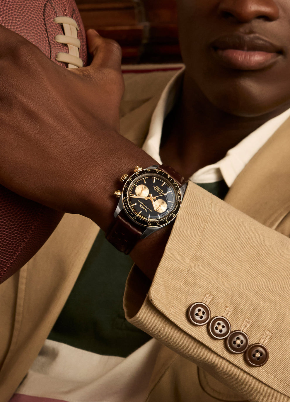 A close-up of a model wearing the Timex Waterbury Heritage Chronograph while holding a football. The watch’s gold subdials, black dial, and brown leather strap stand out against a beige blazer and striped polo, emphasizing a sophisticated, sporty look.