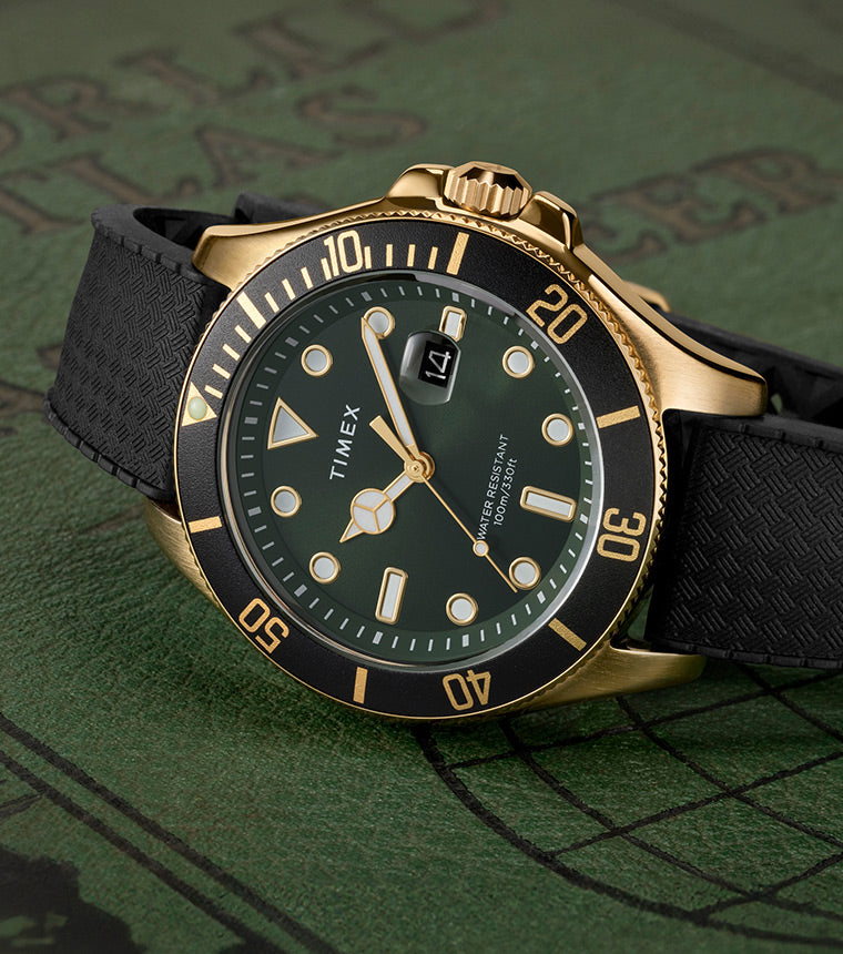 A Timex Harborside Coast watch with a green dial, gold-tone case, and black textured silicone strap rests on an antique-style world atlas cover. The black rotating bezel features gold markers, and the dial includes luminous hands and indices, with a magnified date window at 3 o’clock.