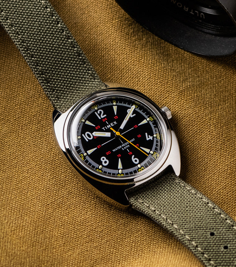 A Timex x Bespoke Post watch featuring a black dial, yellow seconds hand, and olive green fabric strap, displayed flat on a brown textured fabric surface with a camera lens partially visible in the frame.