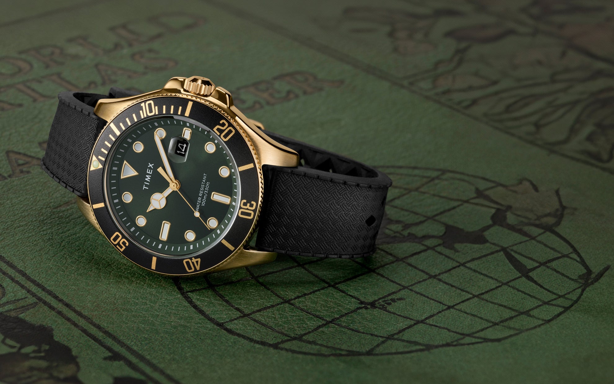 A Timex Harborside Coast watch with a green dial, gold-tone case, and black textured silicone strap rests on an antique-style world atlas cover. The black rotating bezel features gold markers, and the dial includes luminous hands and indices, with a magnified date window at 3 o’clock.