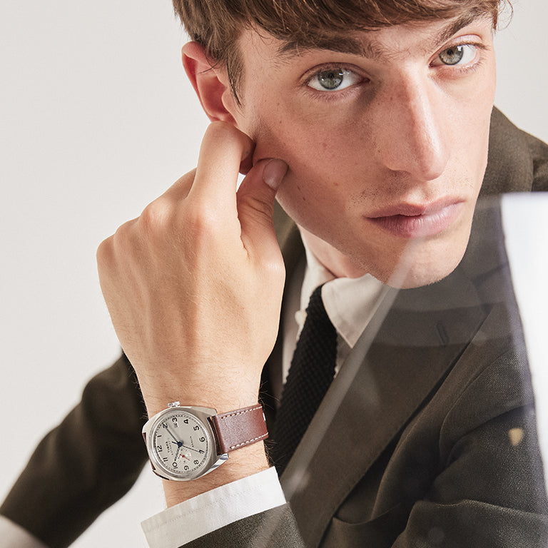 A young man dressed in a dark olive suit, white shirt, and black tie poses with his face resting against his hand. He is wearing a stylish wristwatch with a round silver case, white dial, and brown leather strap. The watch features a clean analog display with black numerals and a blue second hand. The scene is softly lit and minimalist, highlighting the watch as the focal accessory.