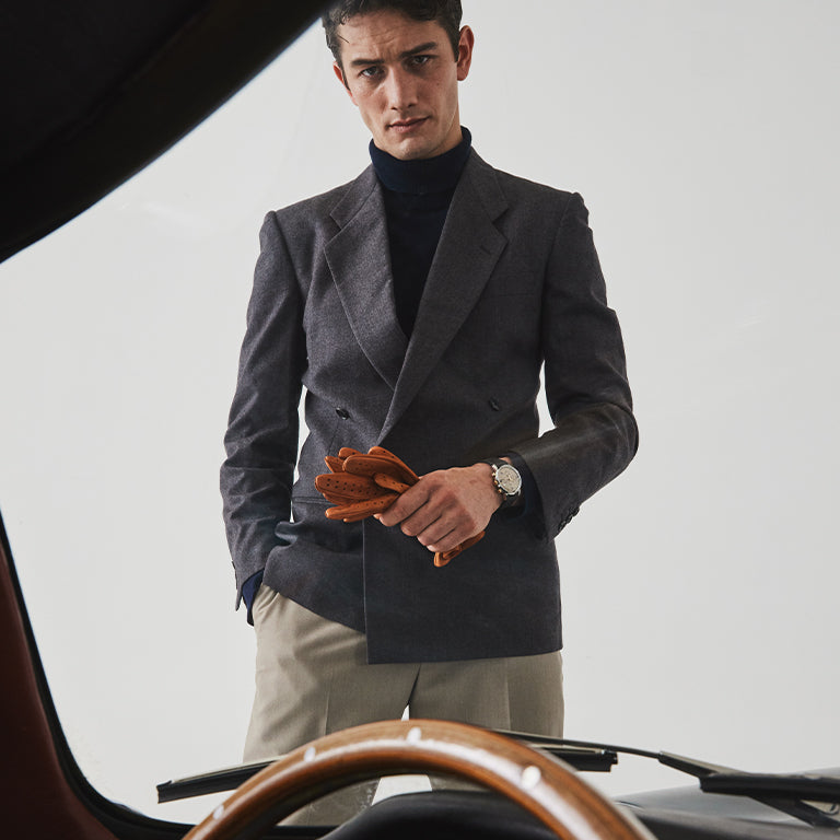 A stylish man stands in front of a vintage car, viewed partially from behind the steering wheel. He wears a tailored charcoal double-breasted blazer over a navy turtleneck, paired with beige trousers. In his hands, he holds a pair of tan leather driving gloves. A classic wristwatch with a brown leather strap and silver case is visible on his left wrist, contributing to the elegant, retro-inspired aesthetic.