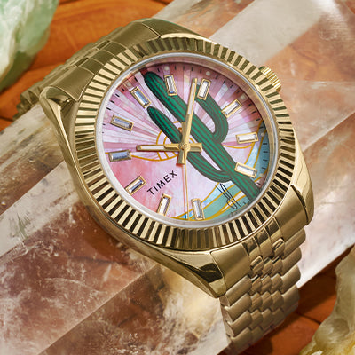 A gold-tone Timex wristwatch with a fluted bezel and link bracelet, displayed diagonally atop a translucent mineral crystal. The watch face features a vibrant desert-themed design with a large green cactus in the center, sunbeam-like pink and white background, and stylized golden hour markers. The Timex logo is positioned on the lower left quadrant of the dial.