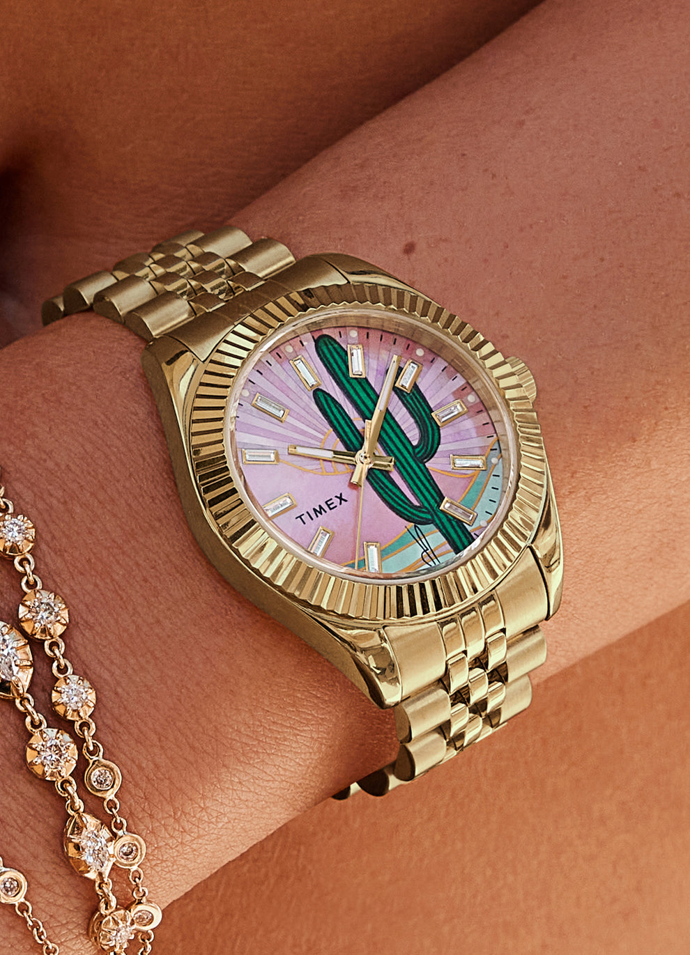 Timex x Jacquie Aiche gold-tone wristwatch worn on a wrist next to layered gold and diamond bracelets. The dial showcases a vivid cactus illustration over a pink-toned background with radiant gold hour markers and a sunburst pattern, set in a fluted bezel and metal link band.