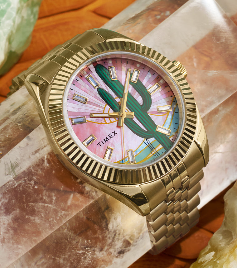 Close-up of a Timex x Jacquie Aiche gold-tone wristwatch placed on a rose quartz crystal, surrounded by other mineral stones. The watch face features a green cactus illustration against a pink mother-of-pearl dial with gold-tone hour markers and hands, encased in a fluted bezel and gold metal bracelet.