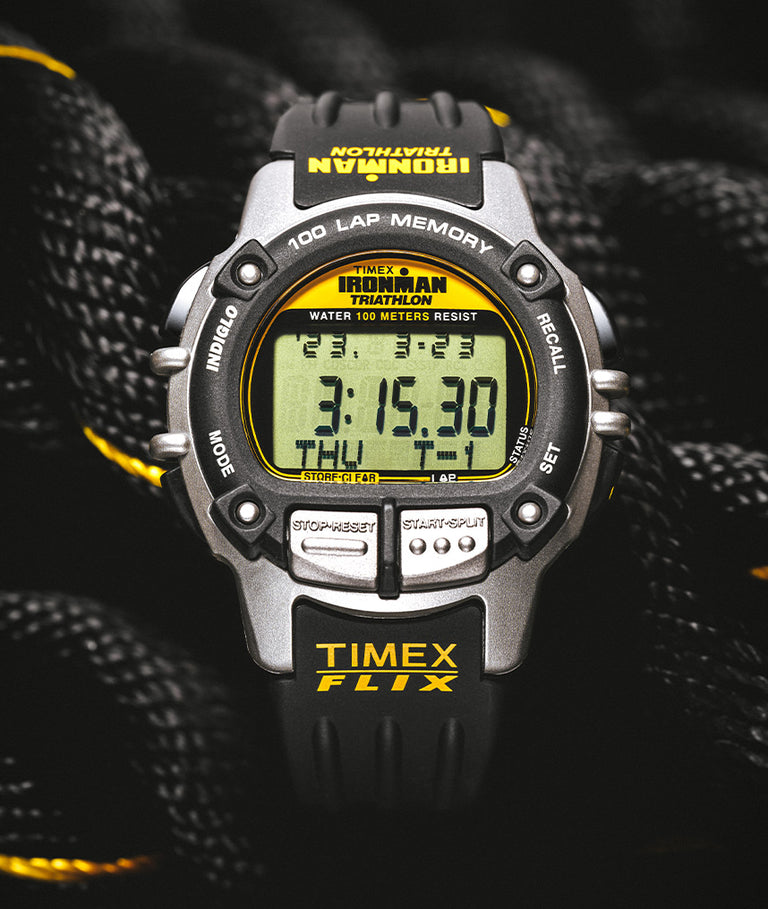 A close-up digital photograph of a Timex Ironman Triathlon digital sports watch with a rugged design, featuring a black and silver case with yellow accents. The watch displays the time as 3:15:30, the day as Thursday, and additional stopwatch and lap memory features. The band is black with "TIMEX FLIX" printed in yellow. The watch is placed on a textured black rope with yellow threads woven through it, complementing the watch’s color scheme.