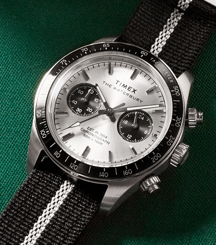 Close-up lifestyle image of the Timex Waterbury Heritage Chronograph with a silver sunray dial, black sub-dials, black tachymeter bezel, and a black fabric strap with a central white stripe, placed on a green felt surface near a cue ball.
