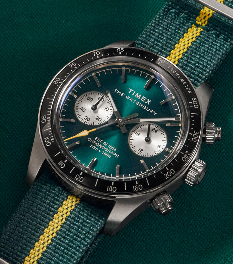 A Timex Waterbury chronograph wristwatch with a vibrant green sunburst dial, silver sub-dials, and white tachymeter markings on a black bezel. The watch features baton hour markers, silver hands with a yellow-tipped chronograph second hand, and is paired with a green and yellow-striped fabric NATO strap. It is laid on a green cloth next to a wooden tray and a red-striped billiard ball.