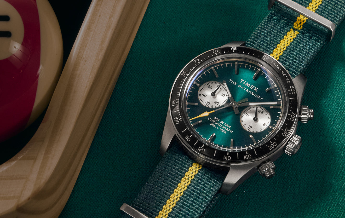 A Timex Waterbury chronograph wristwatch with a vibrant green sunburst dial, silver sub-dials, and white tachymeter markings on a black bezel. The watch features baton hour markers, silver hands with a yellow-tipped chronograph second hand, and is paired with a green and yellow-striped fabric NATO strap. It is laid on a green cloth next to a wooden tray and a red-striped billiard ball.