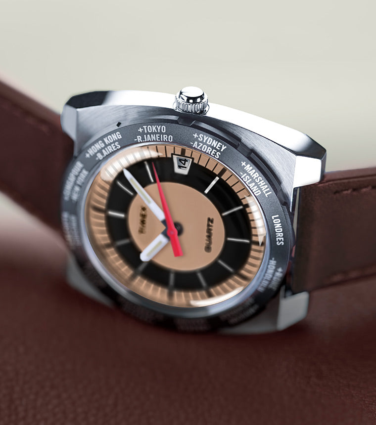 Angled close-up of the Q Timex® 1972 World Time watch resting on a brown leather surface. The stainless-steel cushion case, world time bezel with global city names, and bold black, bronze, and tan dial are in focus, with red, white, and black hands pointing toward the time.