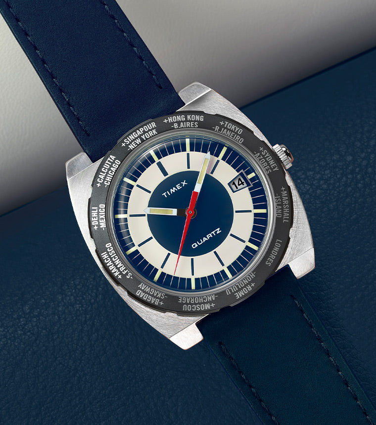 Angled close-up of the Q Timex® 1972 World Time watch placed on a dark blue leather surface. The stainless-steel case and rotating world time bezel frame a blue and cream dial with bold hour markers, red seconds hand, and date window at 3 o’clock. The watch is attached to a navy leather strap with matching stitching.
