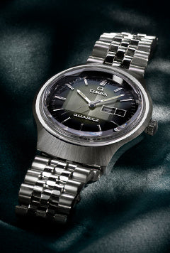 A detailed product image of a Timex Reissue watch with a brushed stainless steel case and a matching bracelet. The black dial features silver hour markers, quartz branding, and a retro day-date window. The watch rests on a dark, silky background.