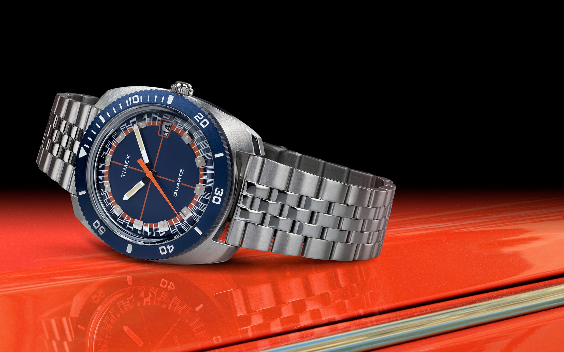 Hero side-angle shot of the Q Timex® 1971 Velocity placed on an orange racecar hood, capturing the reflection of the watch. The brushed stainless case and linked bracelet complement the vivid blue bezel and retro race-day look.