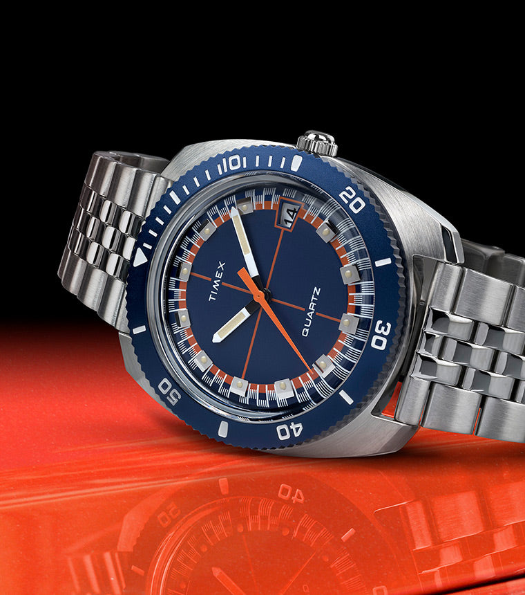 Hero side-angle shot of the Q Timex® 1971 Velocity placed on an orange racecar hood, capturing the reflection of the watch. The brushed stainless case and linked bracelet complement the vivid blue bezel and retro race-day look.