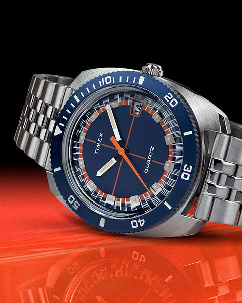 A Timex wristwatch with a brushed stainless steel case and bracelet, featuring a deep navy blue dial and bezel. The watch face includes bold white indices, orange accents on the hands and minute track, and a date window at the 3 o'clock position. The bezel is marked with white numerals in a 60-minute scale. The watch is tilted at an angle, resting on a glossy red surface with a black-to-red gradient background, highlighting its vintage-inspired design.
