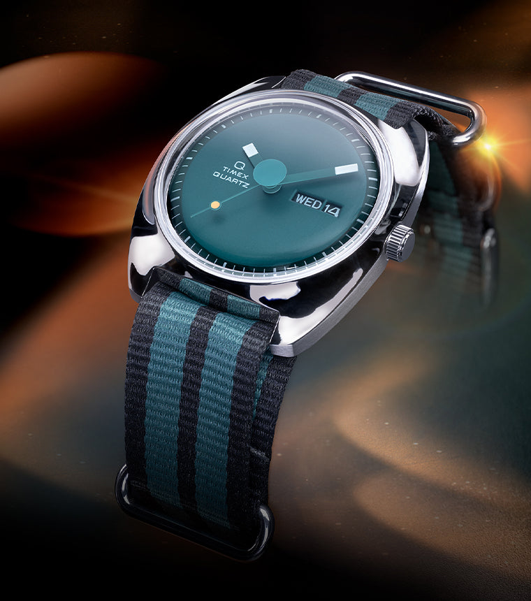 Stylized lifestyle image of the Q Timex® 1975 Enigma watch with a green dial and striped black-and-green NATO strap, illuminated by ambient orange and teal lighting against a cosmic-style background.