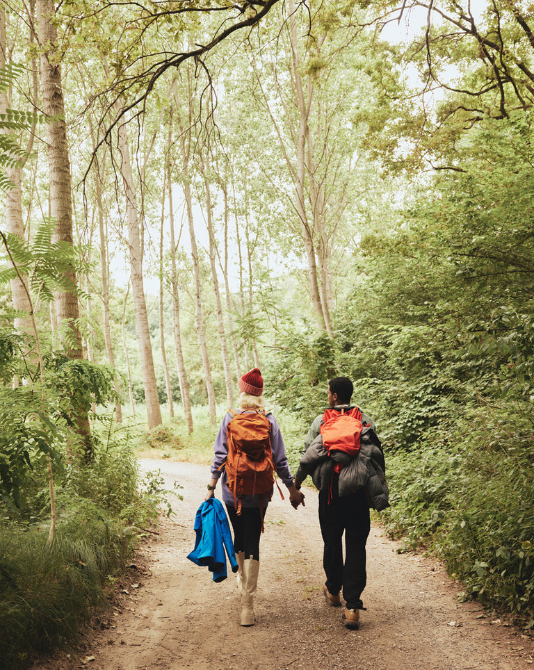 Two hikers, a man and a woman, walk hand-in-hand along a wooded trail surrounded by lush green trees. Both carry large backpacks with outdoor gear; the woman wears light boots, a red beanie, and holds a blue jacket, while the man is dressed in dark clothing with a bright orange pack attached to his bag. The atmosphere is serene and shaded by tall trees, capturing a peaceful moment in nature.