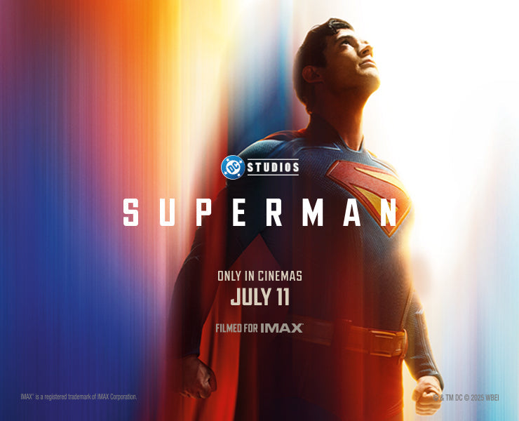 DC Studios’ Superman, featuring the superhero in a blue textured suit with the iconic red and yellow “S” emblem on his chest. He stands confidently with arms crossed, looking upward with a slight smile, illuminated by a bright light from behind. The background is a blend of blue and red streaks, with text reading “SUPERMAN” and “Only in cinemas July 11 – Filmed for IMAX.