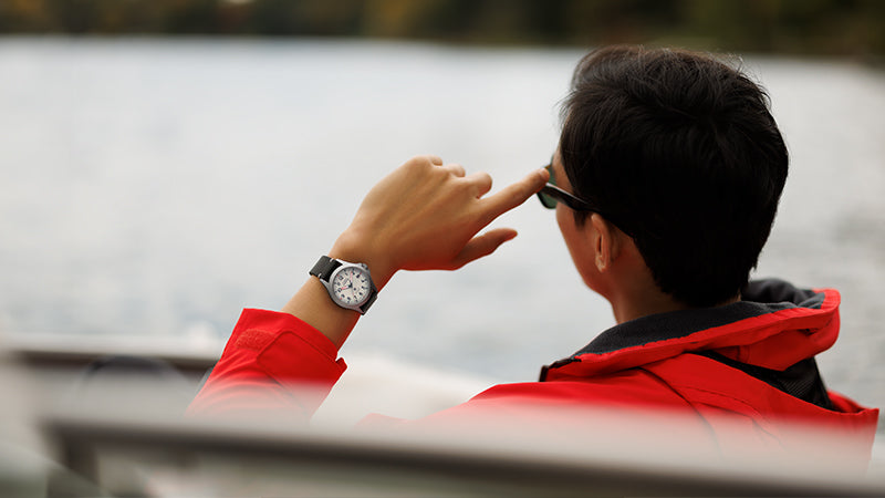 A person wearing a red jacket sits in a boat, adjusting their sunglasses with their left hand, which sports a Timex Expedition watch with a white dial and black strap. The water and distant treeline are softly blurred in the background.