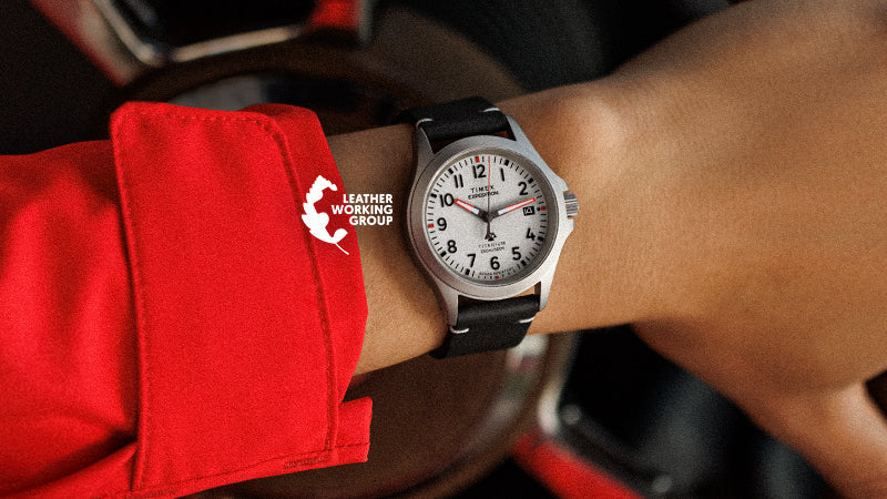 Close-up of a Timex Expedition watch on a wrist, framed by a red cuff, with the Leather Working Group logo overlaid. The white dial, bold black numerals, and date window are clearly visible against a background of a car interior.