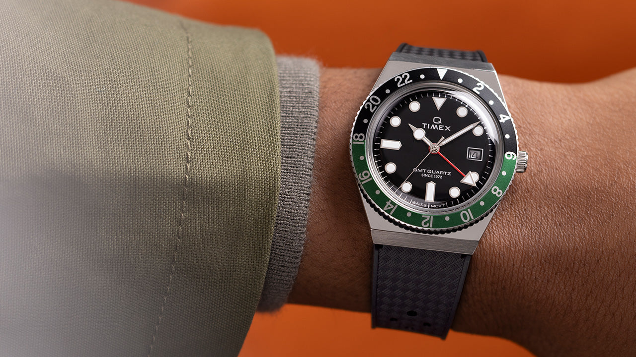 Close-up image of a Q Timex GMT wristwatch worn on the left wrist, featuring a black dial with bold white hour markers, a red GMT hand, and a date window at 3 o’clock. The bezel is split in black and green, marked with 24-hour numerals, emphasizing the dual time zone functionality. The watch is paired with a textured black strap and set against a vibrant orange background, highlighting the retro-sporty design.